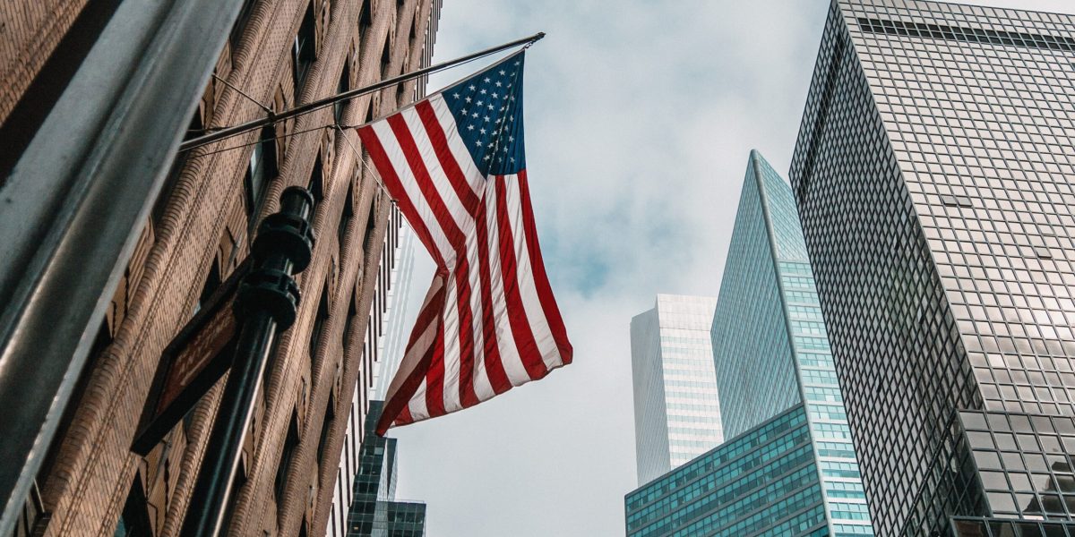 A low angle shot of the USA or United States of America flag on a flagpole near skyscrapers under a cloudy sky